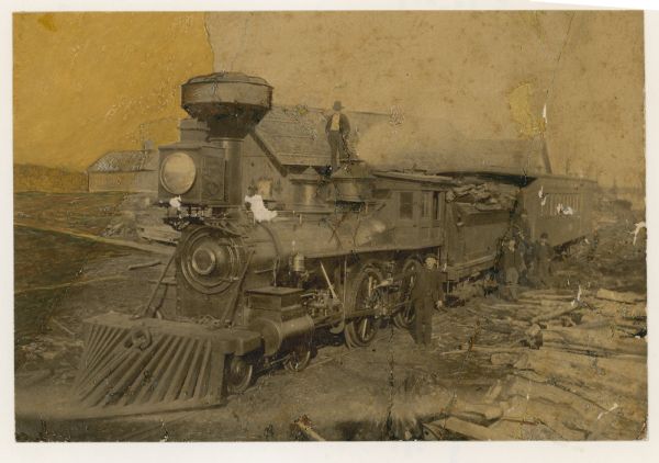 Slightly elevated three-quarter view from front left of a Chicago, Milwaukee, and St. Paul Railway locomotive at Hiles Lumber Company. One man is standing on top of the locomotive, and several men are standing beside it on the right.