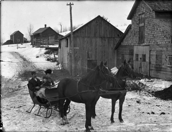 A man, woman, and child posing while seated in a horse-drawn sleigh on a snowy road. Behind them are buildings, a fence, and what may be ice harvesting equipment. The building in the center appears to be filled with hay, with a sign, partly obscured by a telephone pole, that reads: Reme[?] [?]rless Plow.