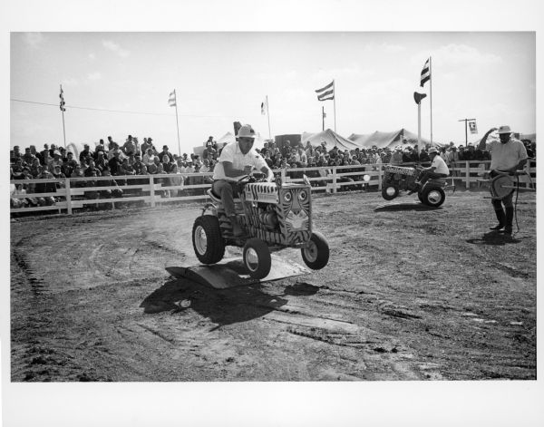 Two Cub Cadet tractors, painted like tigers, perform in a dirt ring as part of a "Wild Animal Act." An actor playing an animal trainer is standing with a chair and a whip, and an audience watches from behind a fence.