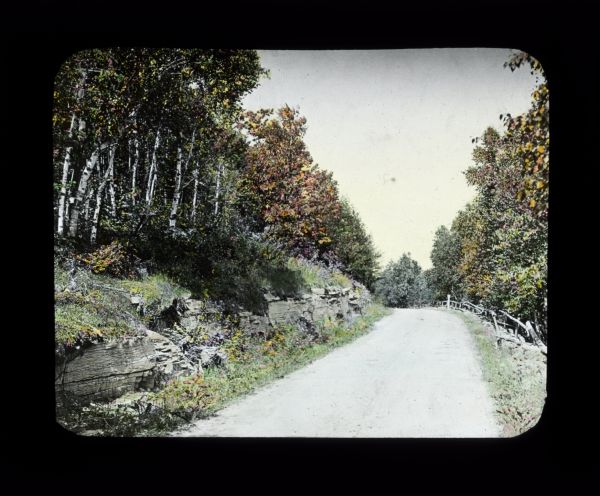 View down rural road lined with trees and rocks. The leaves have turned to autumn colors. There is a fence along the road on the right.