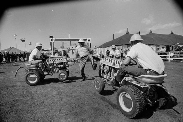 Close-up of two men riding Cub Cadet tractors which are painted like tigers, circling around another man playing an animal trainer and holding a whip in a performance of a "Wild Animal Act." An audience watches from behind a fence in the background.