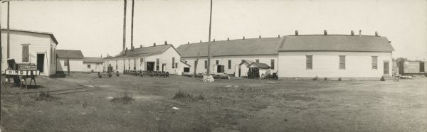 Panoramic photographic postcard view of a railroad depot, with assorted buildings, with barrels on the top of the roofs. Signs on sawhorses on the left read: "O'CALLAGHAN" and "BIRD." There are a number of sets of railroad car wheels on axles in the yard, and on the far right are railroad cars on tracks.