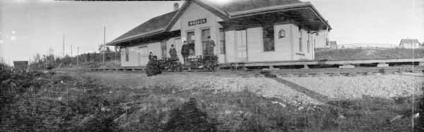 Panoramic view of four men and a child standing in front of the station of the Wisconsin & Michigan Railroad. One of the men is crouching near two handcars or draisines on the railroad tracks. Behind the station are fences and buildings.