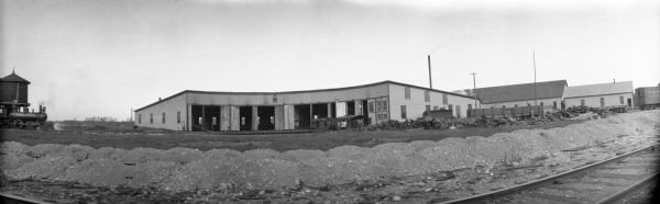Panoramic view of a railroad roundhouse, with railroad tracks and piles of gravel in the foreground. A man is working near a woodpile at the corner of the roundhouse. On the far left a locomotive is moving along the tracks near a water tower.