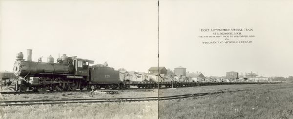 Panoramic right side view of the Dort Automobile special train enroute from Flint, Michigan to Minneapolis, Minnesota via Wisconsin & Michigan Railroad. A man is looking out of the window of the locomotive. Canvas covers with the name "DORT" printed on them cover the automobiles which are on open flatbed cars. Behind the train are industrial buildings and a water tower.