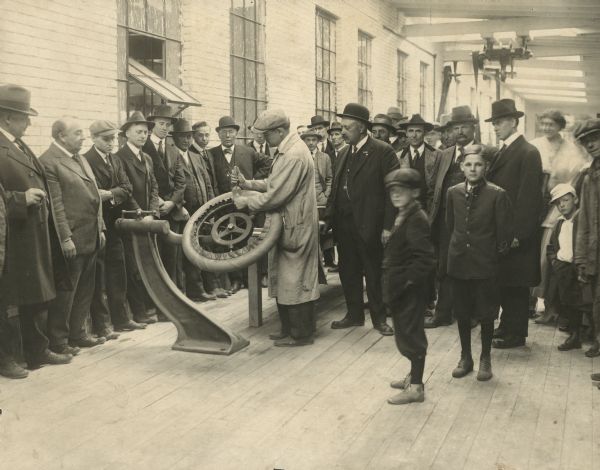 A group of men, women and children are standing and watching as a Gillette employee works on the first tire produced by the Gillette Rubber Company. From left to right are: Mr. Gillette, Hodge Smith, R.W. Hutchens, H. Hutchens, Hans Olson, and Edward Bostwick. The boy in the livery coat is Earl Mertes.