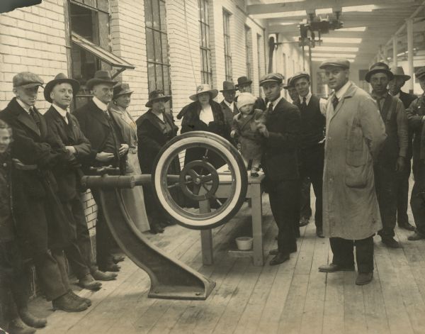 A group of men, women and children standing around the first tire produced by the Gillette Rubber Company. Hodge Smith is standing a baby on a table behind the tire. From left to right are: George Starrs, Sr., R.W. Hutchens, Hodge Smith, Cecil J. Kaufman, and Curly Lawhorn in the long shop coat. 