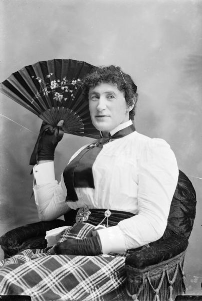 Three-quarter length portrait of an unidentified woman. She is sitting in a wicker chair and is holding an open hand fan behind her head.