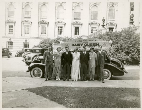 Group portrait of Rose Rosandlich, from Granton, who was elected Dairy Queen in 1935, and her court. They are, from left to right: Harry [?], Bernice Wegner, Henry Dries, Rose Rosandlich, Mr. John Semrod, Mrs. Semrod, and Paul Weiss. Taken outside the Wisconsin State Capitol, which the Queen visited on August 27. The Dairy Queen competition was connected to the Wisconsin State Fair and was sponsored by the Wisconsin Co-op Milk Pool.