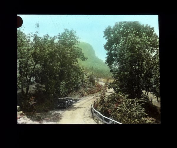 Elevated view of a dirt road between bushes and trees. An automobile is parked on the left side of the road, and a fence runs along the right side. There is a bluff in the distance.