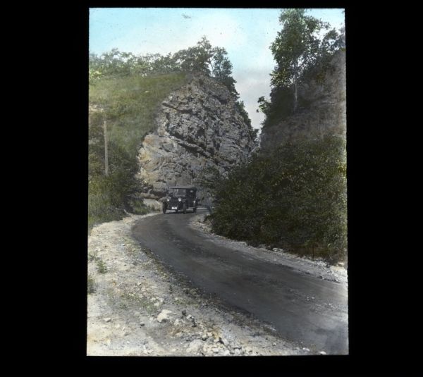View from roadside of an automobile driving up a curve in a road through a large rock formation cut through a bluff.
