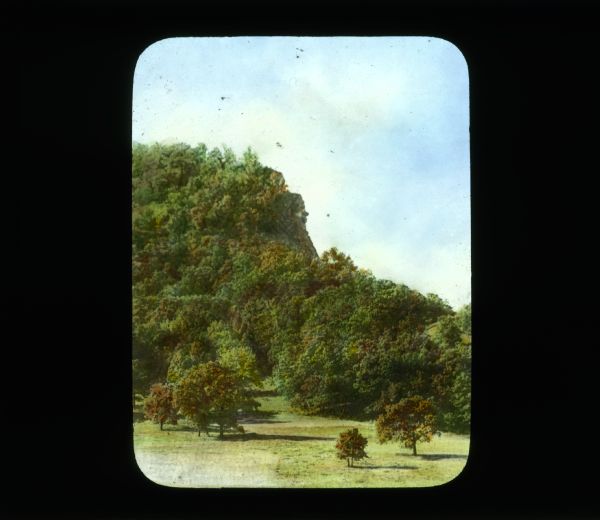 View across field towards bluffs covered with trees. The edge of the bluff has an exposed rock face. The leaves on the trees are autumn colored.