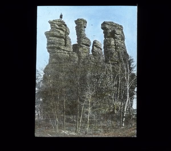 View of four natural rock towers. There is a person (probably a man) standing on top of the one on the far left.