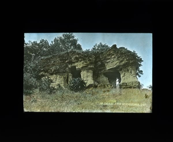 View uphill towards the entrance to the McSherry Cave. A woman is standing at the entrance. Trees and shrubs are growing on top of the cave.