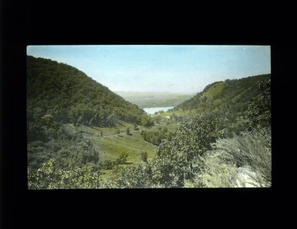Elevated view of a valley between tree-covered hills. There is a lake or river, and a far shoreline in the distance.
