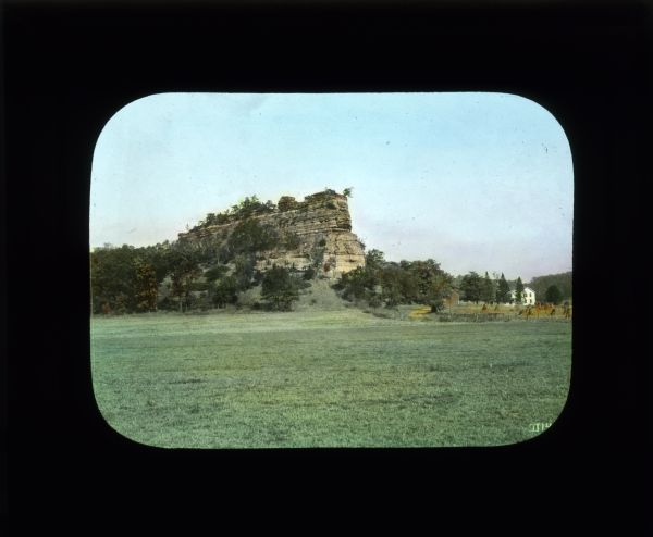 View across a field towards a tall bluff. On the right is a house and barn at the base of the rock formation.