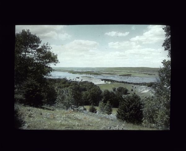 Elevated view from a hill of a lake or river surrounded by fields and hills. The water is either marshy, or has been flooded, as trees and plants are growing in the water. Near the lake or river is a cone-shaped mound below the hill in the foreground.