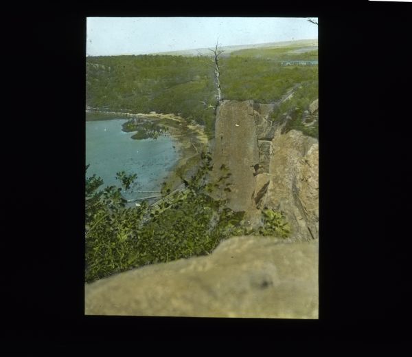 View from a rock cliff looking down on a lake with a shoreline, surrounded by tree-covered hills. The shoreline is sandy and marshy. Directly below the cliff in the foreground are railroad tracks.  This appears to be Devil's Lake    