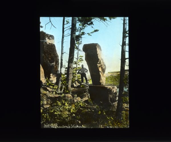 View through trees towards a man posing next to a large rock formation of a balancing rock. On the right is another rock formation. Below in the background is a lake. Caption reads: "Balanced Rock, Devil's Lake."