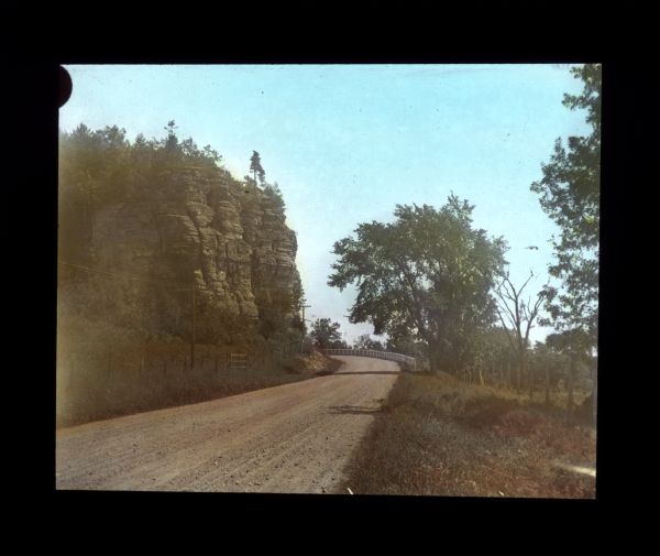 View across unpaved rural road that curves around to the left around a large rock formation with an exposed rock face with trees growing on the top. There is a fence and power lines on the left along the road, and on the right near the curve in the road is a guard rail, and a fence in a field.