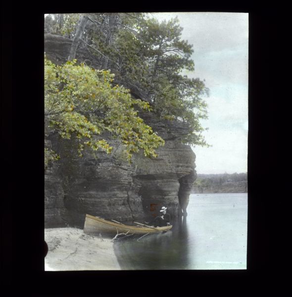 A man relaxing in a canoe on a river or lake. Behind him is a rock formation covered with trees.