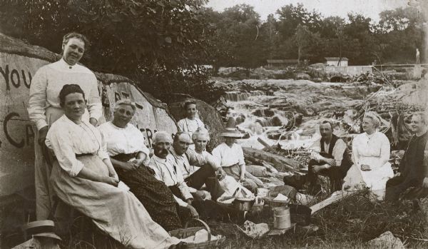 Group portrait of people picnicking on the shoreline of a river. In the background is an unidentified waterfall, with tree limbs and branches stuck in the rocks and along the shoreline. On the left beside the group is a painted sign on the side of a rock, which reads, in part: You [?]e ___ at- Ca[?] [?]er[?]. In the background a man is walking on a small bridge over the falls.