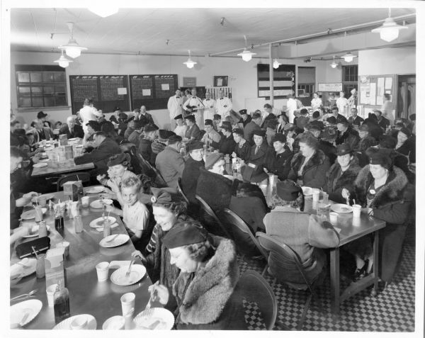 Slightly elevated view of a band playing and singing in a crowded cafeteria. The cafeteria is not identified, but signs on the walls advertise Gargoyle Coffee and Eilers Griddle Cake Service.