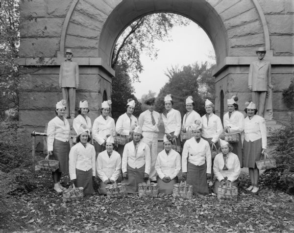 Group portrait of a group of Oscar Mayer canvassers posing in front of the Camp Randall Memorial Arch. They are all wearing uniforms and holding baskets of Oscar Mayer Wieners cans. One man is standing in the center.