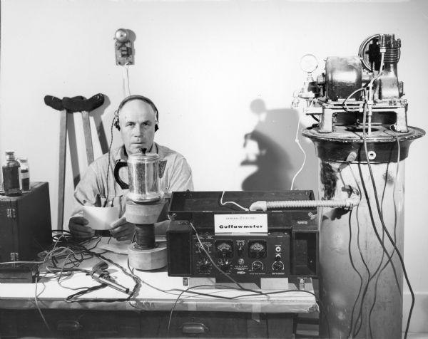 A man wearing headphones is sitting behind a table with the Guffawmeter. The caption reads: "Guffawmeter constructed by Paul Vanderbilt for the State Historical Society's Joke Collecting Project. The meter at this reading is at .017."