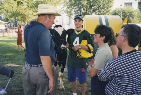 Television weatherman personality Willard Scott at the Smithsonian Folklife Festival on the National Mall in Washington, DC. Scott broadcast a segment for NBC’s Today show adjacent to Bill Casper’s iconic Green Bay Packers ice shanty. Scott is talking to a group of people, and a young man wearing a Packer's jersey is standing and holding the bridle of a cow. The ice shanty is in the background.