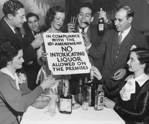 Men and women celebrating the end of Prohibition, around a table with bottles of various alcoholic beverages. They are holding a sign that reads: "In Compliance With The 18th Amendment, No Intoxicating Liquor Allowed On The Premises." Four of the people are setting fire to four corners of the sign.