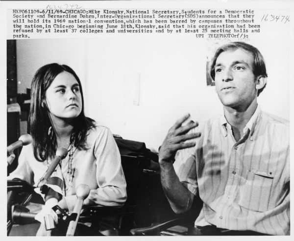 Still from the film "Underground" by Emile de Antonio. Mike Klonsky and Bernardine Dohrn are at a press conference, sitting behind microphones. Dohrn is sitting on the left, looking at Klonsky speaking on the right.