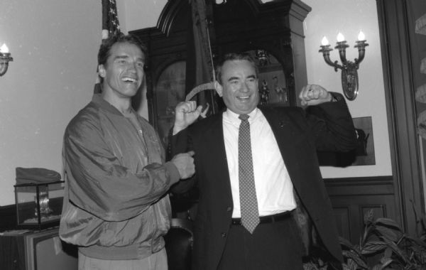 Indoor portrait of actor Arnold Schwarzenegger posing with Wisconsin Governor Tommy Thompson, at the Wisconsin State Capitol. Schwarzenegger had come to Wisconsin as chairman of the President's Council on Physical Fitness. Governor Thompson is flexing his arms and Arnold Schwarzenegger is pointing to him. Both men are smiling.
