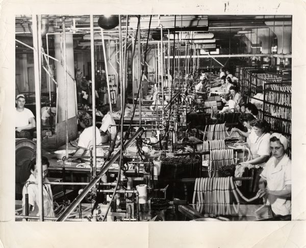 Elevated view of about 29 employees manufacturing sausages at the Oscar Mayer & Co. plant. Caption reads: "This view of the sparkling Sausage Kitchen at Oscar Mayer & Co., Madison, shows Yellow Band wieners in production. These highly productive and skilled employees work as a team, to produce an average of more than 50,000 pounds of wieners daily."