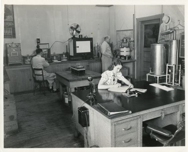 Two man and one woman are working in the laboratory of the Oscar Mayer Company plant. In the foreground the woman is writing in a book at a desk with a telephone, and behind her two men are looking at machines.