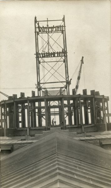 View along top of roof towards the fourth State Capitol building under construction, with erection tower and gin pole behind the girders of the dome substructure. At least five men are working among the girders, and a few men are standing on the top of the erection tower. Caption on back reads: "Dome proper looking over and taken from the roof of West Wing. Shows dome up to 106' lever [sic] and also shows erection tower in center of dome being extended on up to 240' level. Gang all ready to fleet gin pole up to next elevation."