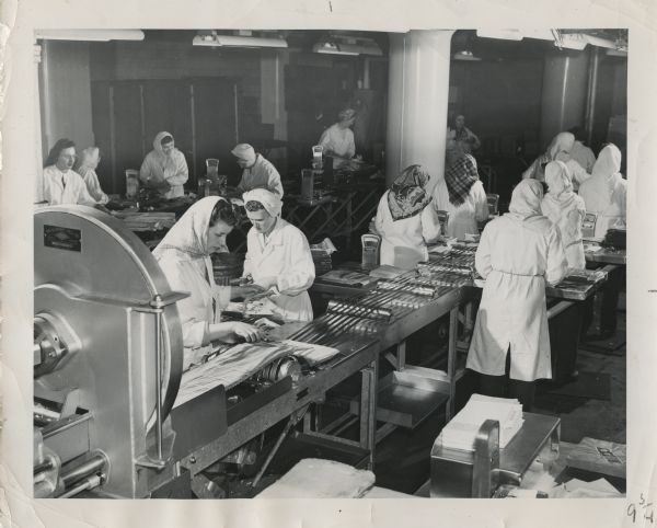 Women preparing sliced bacon at the Oscar Mayer & Co. plant in Madison. At least fifteen women are working, most most of them wearing headscarves. Caption reads: "One of the most modern production lines at Oscar Mayer & Co. is in the Sliced Bacon Department. As the bacon sides pass through the high speed slicers they are weighed into one pound packages and packed for shipping. A great deal of care is exercised by the women in weighing and sorting bacon for the Yellow Band package."