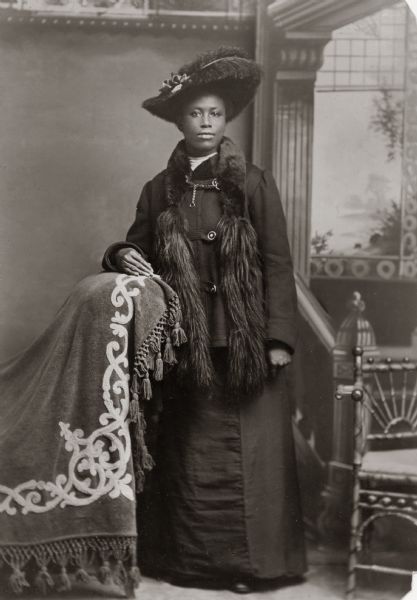 Studio portrait of Mrs. William W. "Fannie" Hendricks in formal dress, including a fur scarf and feather hat, in front of a painted backdrop.