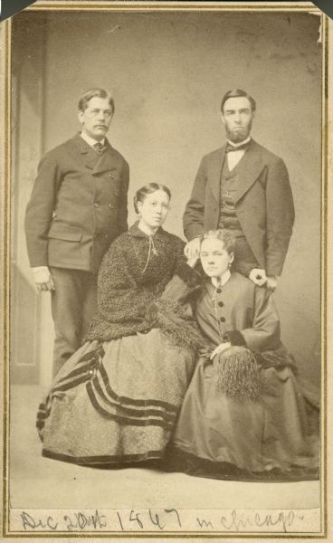 Formal studio group portrait in front of a painted backdrop of William Upham (standing, left), his wife Mary Upham (seated, left), and a man and woman, both unidentified. Both women are holding hats with ostrich feathers in their laps.