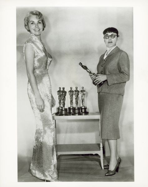 Janet Leigh and Edith Head standing next to a table with five Oscar statues. Edith is holding an Oscar statue in her hands. Janet is wearing a full-length gown Edith designed for her to wear to the Oscar ceremony.