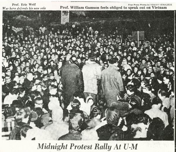 Elevated view over tightly packed crowd towards Philosophy Professor Frithjof Bergmann, shown from the back standing at a podium, calling for a United States withdrawal from Vietnam at a teach-in rally held at the University of Michigan. Two other men are standing on either side of him, and they are surrounded by about 3,000 students and faculty. 