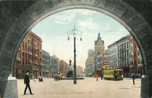 View through a City Hall archway of the city square, which includes a statue, a streetcar, and a decorative lamp post. Caption reads: "City Hall Square, Milwaukee, Wis."