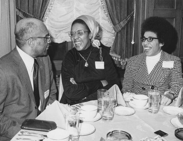 Group portrait of three people seated at a dining table. Wesley Scott, executive director of the Milwaukee Urban League, is facing Geraldine Wilson, who was active with the Head Start program in Milwaukee, and Gwendolyn Gilbert. Both women are laughing.