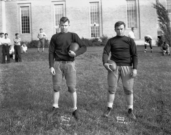 Two Madison Edgewood High School football players posing in uniform outdoors. Vic Ripp, all-city halfback (L) and Fonse Hughes, fullback (R). The Edgewood High School building is behind them. In the background other young men are sitting and standing on the lawn.
