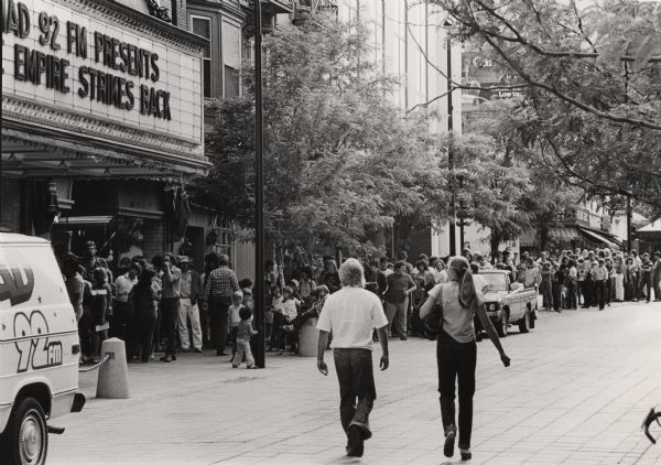 View down street towards a large crowd gathering in a line outside the Orpheum Theatre on State Street for a free 10AM Madison premiere of <i>The Empire Strikes Back</i> sponsored in part by WMAD-FM. A radio station van is on the left.
