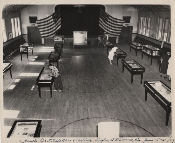 Elevated view of people examining display cases in a gymnasium containing objects from the French Gratitude Train. Two U.S. flags are hanging at the far end of the room flanking a stage, above which is a  folded up basketball hoop.