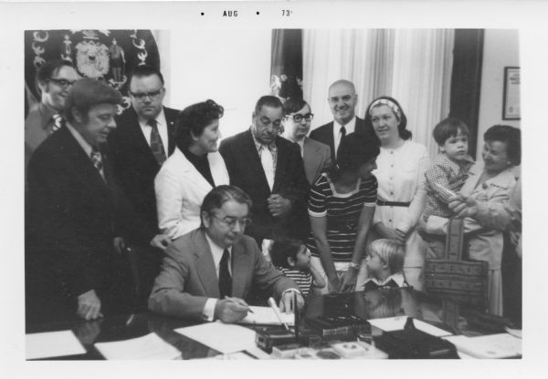 Governor Pat Lucey signing Chapter 115 ensuring public education for children with disabilities as parents, children and others look on. Midge Miller stands directly behind the Governor.