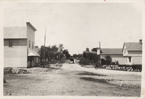 Postcard view of a street in Egg Harbor. A steam roller and pedestrians are on the road. Several buildings line both sides of the street. Caption reads: "St. Scene, Egg Harbor, Wis."