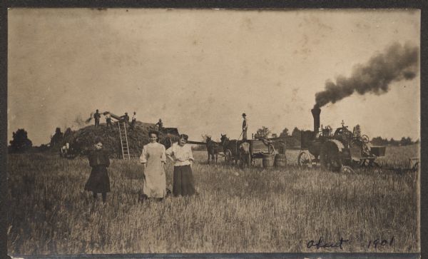 Three women, identified as Louisa, Esther, and Engeline, standing in a field (possibly soybean) on the Julian Corbisier farm. Men in the background are threshing with a belt-driven steam powered tractor.