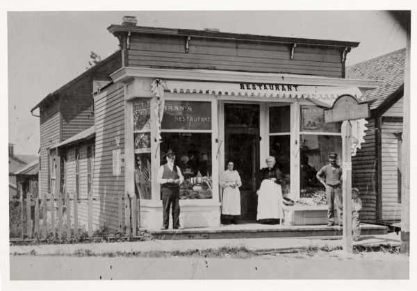 James R. Mann, and Eleanor Mann, proprietors of Mann's restaurant, standing in front of their storefront, with the clerk, Joe Haberlie, and an unknown woman and a boy.
